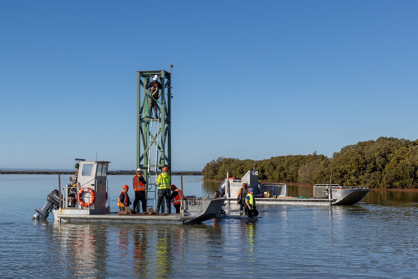 pearl nail in water with two men on board, others in water shallow water in waters installing a white pole