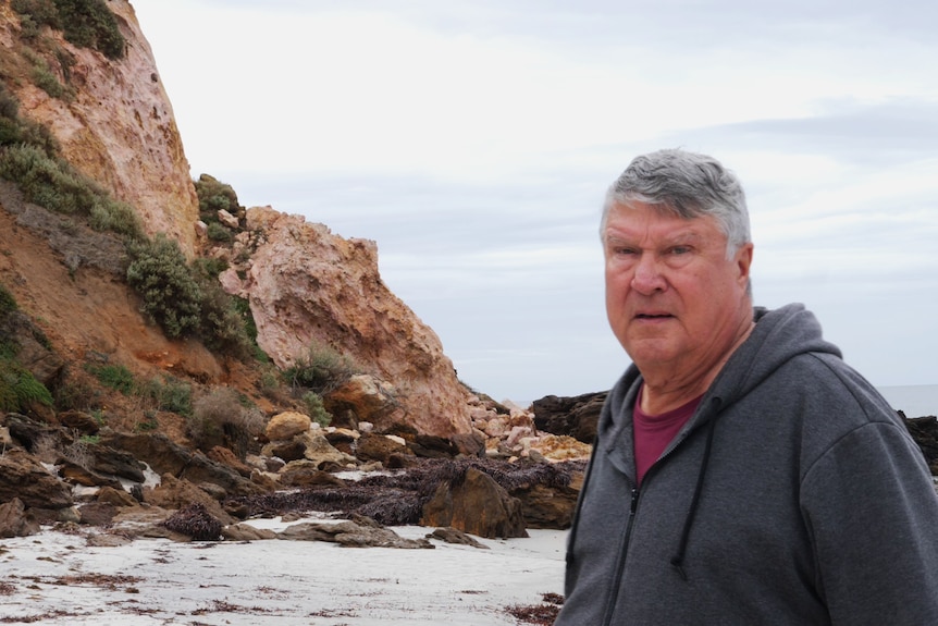 Man in foreground looking at camera, standing next to crumbling rock, ocean and rocks in background